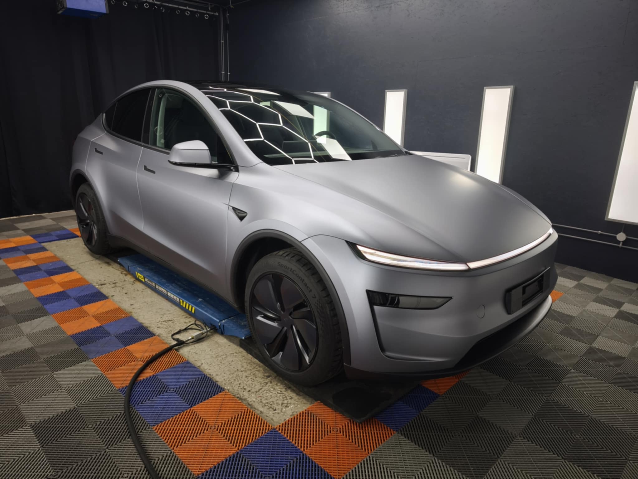 Voiture électrique gris mat dans un atelier, posée sur un lift; film de carrosserie partiellement couvrant le toit avec motif hexagonal, ambiance labo. inconvénients du film ppf voiture intégrés subtilement.