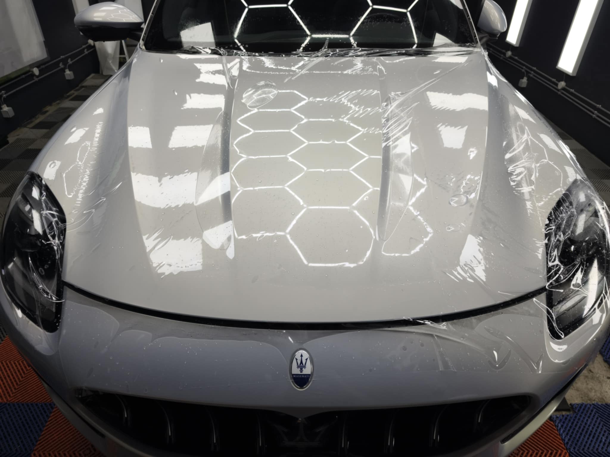 Voiture Maserati gris clair avec film protecteur sur le capot brillant, reflet des lumières en motif hexagonal, affichant les inconvénients du film ppf voiture dans l’illustration.